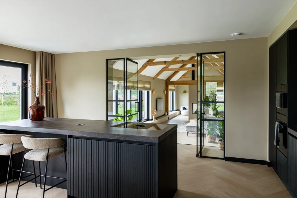 Keuken met stalen taatsdeuren en zichtlijn naar de woonkamer met eiken spanten in de verbouwde jaren 50 boerderij in Lettele.